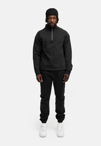 Schwarze Sportbekleidung bestehend aus einem Reißverschluss-Pullover und passenden, taillierten Hosen, kombiniert mit weißen Sneakers. Schlichte, strukturierte Stoffe.