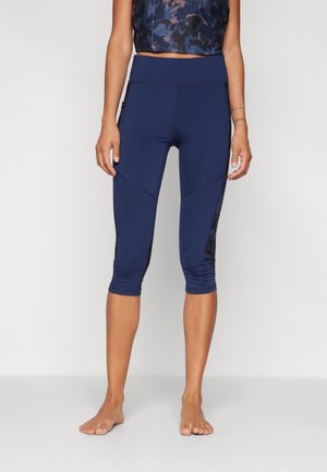 Frau in marineblauen, knielangen Leggings mit gemusterten Seitenteilen, steht barfuß vor einem schlichten, hellen Hintergrund.