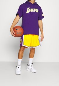 Persona che indossa un cappuccio viola dei Lakers e pantaloni corti gialli da basket dei Lakers, tiene un pallone da basket Nike in una mano, in piedi su uno sfondo semplice.