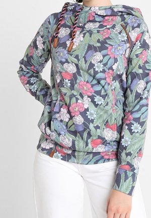 Femme portant un sweat à capuche à manches longues à motif floral avec cordons et un pantalon blanc, debout avec une main sur la hanche.