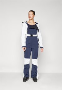 Tumedate sinine ja valge suusakombinesoon, millel on lukuga eesosa, lai krae ja reguleeritav vöökoht. Omab pikki varrukaid ja laienevaid jalgu.