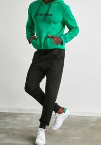 Grön hoodie med "TRÈS BIEN" i svart text, kombinerad med svarta joggarbyxor och vita sneakers med gröna accenter. Avslappnad sportig stil.