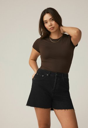 Fiatal nő egyenes sötét hajjal, barna testhezálló pólót és fekete farmer rövidnadrágot visel, amelynek a szegélye foszlott, egyik kezével a nyaka mögött pózol.