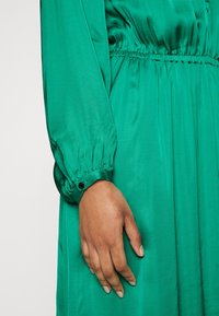 Grön satin klänning med långa ärmar och rynkade manschetter samt smockad midja. Slät textur med en mjuk lyster.