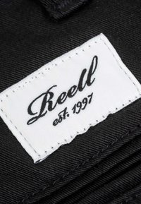 Weißes rechteckiges Stoffetikett mit schwarzem kursivem Text "Reell est. 1997", auf schwarzem strukturiertem Stoff aufgenäht.