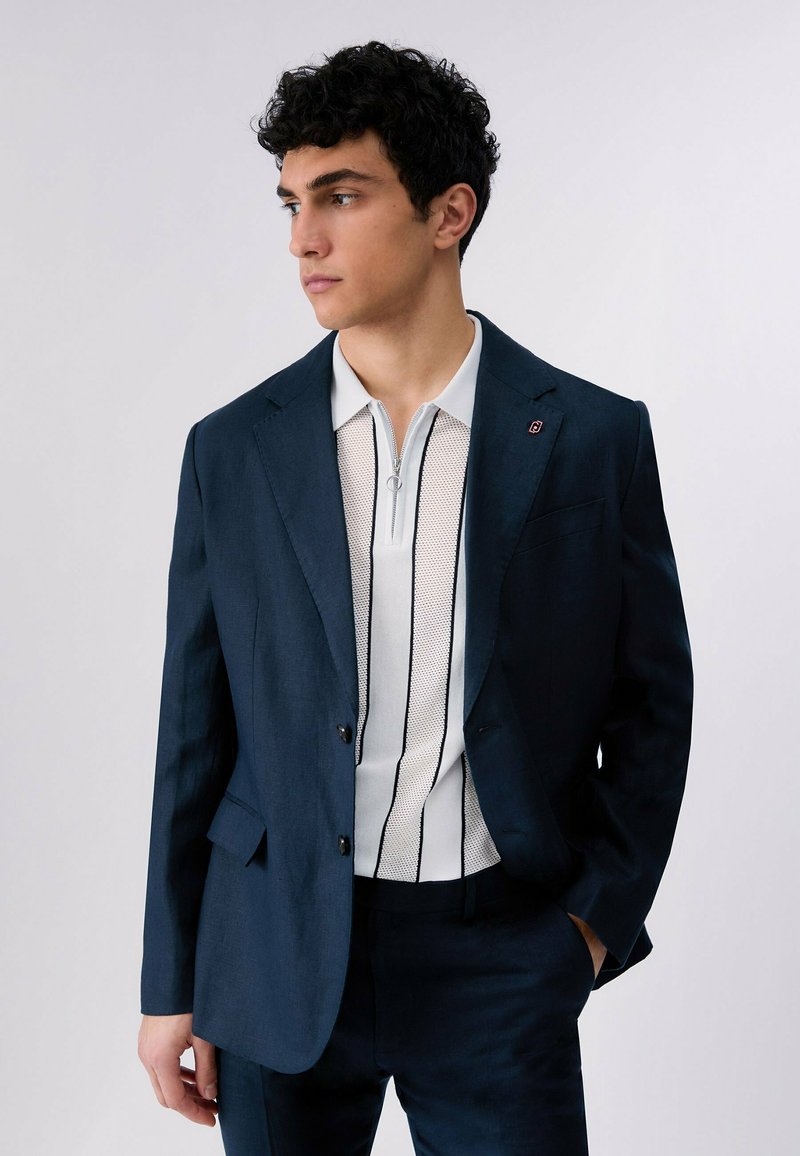 Jeune homme en costume bleu marine et chemise blanche zippée avec des rayures verticales noires, debout avec une main dans la poche, regardant vers la gauche.