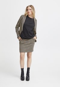 Ensemble blazer et jupe à motif pied-de-poule dans des tons de marron et noir, associé à un haut noir uni. Tissu lisse avec un ajustement sur mesure. Bottines noires à la cheville.