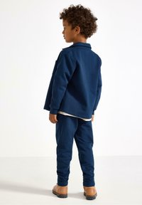 Jong kind met krullend haar, gekleed in een marineblauwe sweatshirt en broek, met tan kleurige laarzen, staand en zijwaarts gezichten op een witte achtergrond.