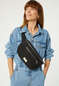 Sac en tissu noir à bandoulière avec deux compartiments zippés et un patch logo, porté sur une veste en denim à manches retroussées.