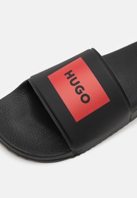 Svart sandaler med glatt gummisåle, med en bred stropp i svart som har en rød rektangulær logo som viser "HUGO" i svart.