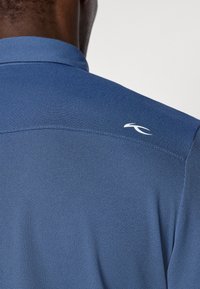 Pogled na osebo v modri polo majici s teksturo, ki ima majhen bel logotip na zgornjem delu hrbta blizu rame.