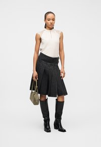 Γυναίκα που φοράει λευκό τοπ χωρίς μανίκια με δαντέλα, μαύρη φούστα με πιέτες, μαύρες μπότες μέχρι το γόνατο και κρατάει μια πράσινη τσάντα χειρός.