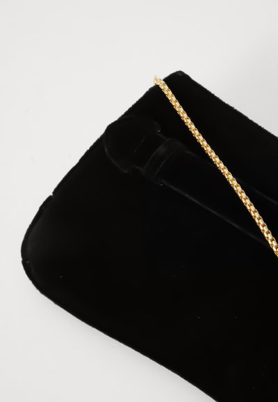Svart fløyel clutch-veske med en gullkjede-rem som hviler på en hvit overflate.