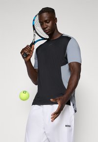 Svart og grå kortermet treningstrøye med teksturerte paneler, kombinert med hvite shorts. En tennisracket og en gul tennisball er til stede.