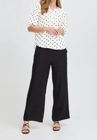 Femme portant un chemisier blanc à pois noirs, un pantalon large noir et des sandales noires à lanières, debout les mains jointes.