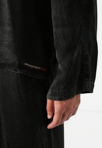 Chemise noire à manches longues avec une texture froissée, présentant une étiquette de logo visible et des coutures détaillées le long des manches et de l'ourlet.