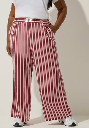 Person iført vidde bukser med lodrette røde og hvide striber, en hvid t-shirt og hvide sneakers, stående med den ene hånd i lommen.