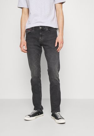 Person trägt verblasste schwarze Slim-Fit-Jeans, ein hellgraues Kurzarmhemd und schwarze Low-Top-Sneaker vor einem schlichten weißen Hintergrund.