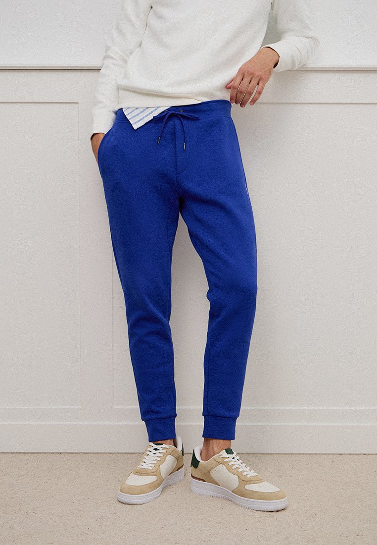 Osoba w jaskrawo niebieskich dresowych spodniach, w beżowych i białych sneakersach, w białej bluzce z długim rękawem, stojąca przed białą panelem ściennym.