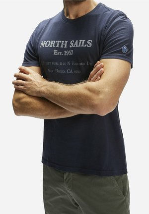 Mężczyzna z założonymi ramionami, ubrany w granatowy t-shirt "North Sails Est. 1957" i oliwkowe spodnie, pokazany od szyi do bioder.
