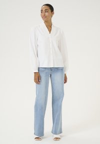 Blouse blanche à manches longues avec un col en V et textured, associée à un jean large bleu clair et des talons ouverts blancs.