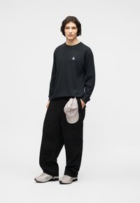 Νεαρό άτομο με μαύρο μακρυμάνικο πουκάμισο με μικρό λογότυπο, άνετο μαύρο cargo παντελόνι, γκρι αθλητικά παπούτσια, κρατώντας ένα ανοιχτό γκρι καπέλο στην τσέπη.