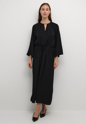 Rochie neagră, cu mâneci lungi, decolteu în V și talie cu șnur. Material neted, croi lejer și lungime până la jumătatea gambei. Tocuri negre ascuțite completează look-ul.