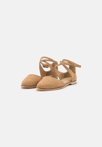 Sandalias planas de ante beige con punta afilada, correas en los tobillos y tacones bajos sobre un fondo claro y liso.