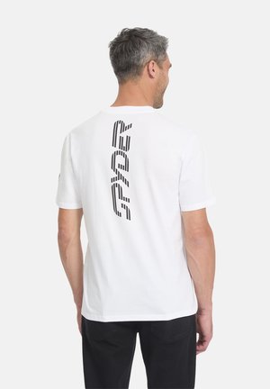 Homme aux cheveux gris portant un T-shirt blanc avec le texte vertical noir "PEPE" dans le dos, associé à un pantalon noir, tourné de dos sur un fond uni.