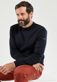 Dunkelblauer Strickpullover mit hohem Kragen, seitlichen Knopfakzenten an der Schulter und gerippten Bündchen. Getragen über rostfarbenen Hosen.