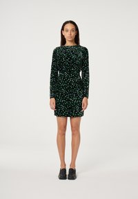 Schwarzes Samtkleid mit grünen und blauen Blattmustern, mit langen Ärmeln und einem drapierten Ausschnitt. Kombiniert mit schwarzen Schlupfschuhen.