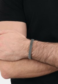 Pulsera tejida negra con un patrón en zigzag, hecha de un material texturizado, llevada en una muñeca con pelo claro, sobre un fondo de camisa oscura.
