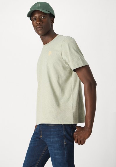 T-shirt en coton vert clair à manches courtes, portant un petit logo de fleur jaune sur la poitrine, associé à un jean bleu foncé.