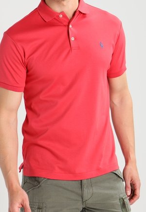 Rotes Polohemd mit Kragen, kurzen Ärmeln und blauem Emblem auf der linken Brust. Der Stoff wirkt glatt und leicht.