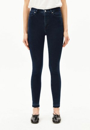 Persoon die donkerblauwe skinny jeans en zwarte ballerina's draagt, staand tegen een witte achtergrond met ontspannen handen langs het lichaam.