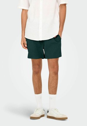 Homme portant une chemise blanche à manches courtes avec boutons, un short texturé vert foncé avec des poches, des chaussettes blanches montantes et des baskets blanches.