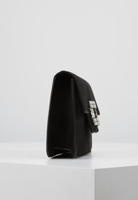 Bolso de mano rectangular negro con tapa y una gran hebilla de strass rectangular, de pie sobre una superficie blanca.