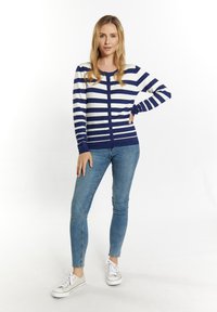 Tamsiai mėlynas ir baltas dryžuotas kardiganas su apykakle, mygtukų uždarymu ir ilgomis rankovėmis, derinamas su mėlynomis siaurėjančiomis kelnėmis ir šviesiais sportiniais batais.