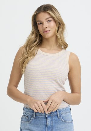 Mladá žena s dlhými blond vlasmi, oblečená v béžovo-modrom pruhovanom tielku bez rukávov a modrých džínsoch, stojaca pred bielym pozadím.