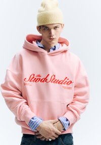 Rózsaszín kapucnis pulóver piros szöveggel, elülső zsebbel, kék csíkos ingre húzva. Világos sárga kötött sapkával kiegészítve.