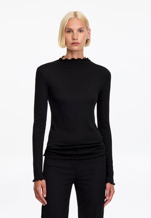 Mujer rubia con el pelo corto, vistiendo una camiseta negra de manga larga con bordes con volantes y pantalones negros, de pie frente a un fondo blanco liso.