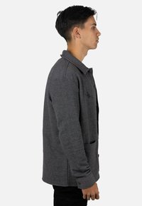 Chaqueta gris con textura, cuello con solapa, cierre con botones y dos bolsillos delanteros. Puños ajustables con un detalle sutil de logo en el pecho.