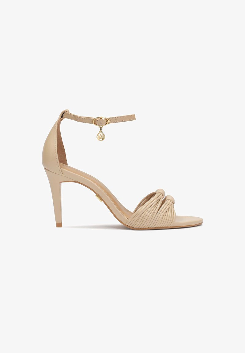 Nude high-heeled sandal featuring a knotted front, smooth texture, adjustable ankle strap with gold hardware, and a slender stiletto heel.