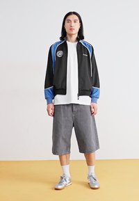 Black and blue zip-up track jacket with round logo, paired with a white t-shirt and gray knee-length shorts. Silver sneakers and white socks.