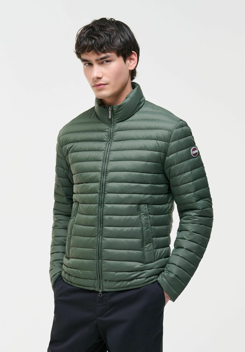 Junger Mann trägt eine grüne, gesteppten Jacke mit Reißverschluss, Stehkragen und seitlichen Taschen, Hände in den Taschen einer marineblauen Hose, schaut nach vorne.