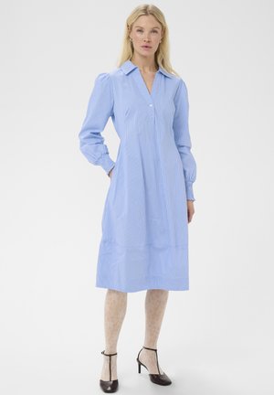 Donna che indossa un abito midi azzurro chiaro a maniche lunghe con colletto e sottili righe verticali, collant beige fantasia e scarpe nere con tacco.