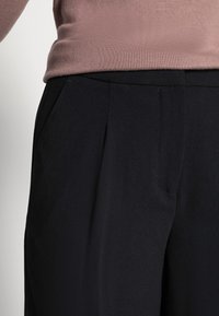 Zwarte broek met plooien, gemaakt van een gladde stof. De tailleband is zichtbaar, met een fijne textuur en een subtiele glans.