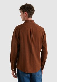 Brązowa koszula o fakturowanym wykończeniu z kołnierzykiem zapinanym na guziki, z plisą z tyłu i mankietami zapinanymi na pojedynczy guzik, w połączeniu z dżinsami, widok z tyłu.