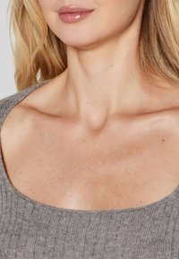 Top grigio a coste con un ampia scollatura curva. Tessuto liscio con sottili dettagli della pelle visibili, incorniciato da capelli sciolti e mossi.