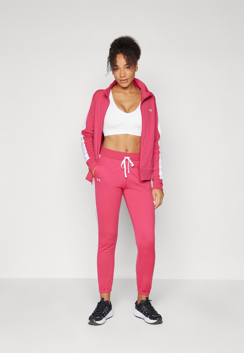 Completo sportivo composto da una giacca zip rosa con strisce bianche, pantaloni jogger rosa coordinati e una maglietta cropped bianca. Le scarpe da ginnastica nere completano il look.